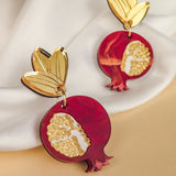Gold and red handcrafted earrings shaped like pomegranates with gold leaf tops, placed on a soft white fabric background, showcase expert craftsmanship and artistry.