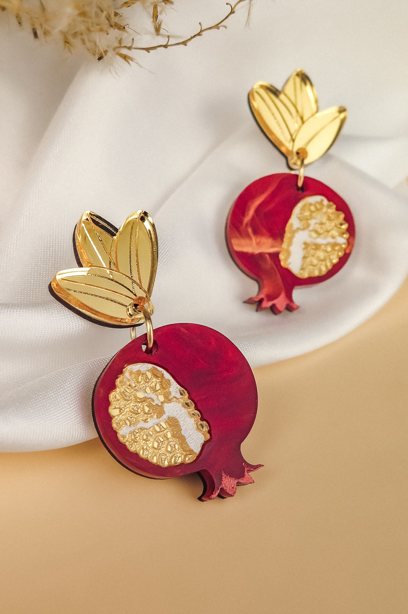 Gold and red handcrafted earrings shaped like pomegranates with gold leaf tops, placed on a soft white fabric background, showcase expert craftsmanship and artistry.