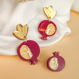 Gold and burgundy handcrafted earrings shaped like pomegranates with leaf-shaped tops and gold detailing, displayed upright on white fabric. A matching pomegranate brooch lies in front on a beige surface, with dried flowers above.
