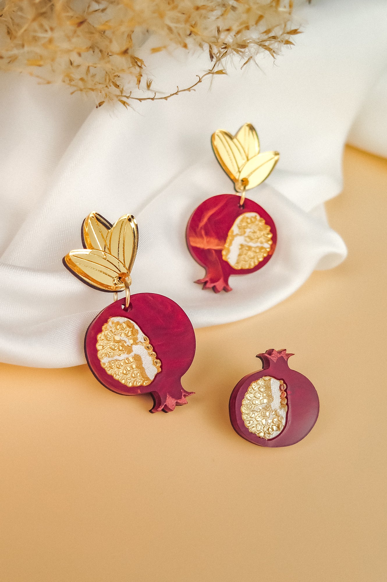 Gold and burgundy handcrafted earrings shaped like pomegranates with leaf-shaped tops and gold detailing, displayed upright on white fabric. A matching pomegranate brooch lies in front on a beige surface, with dried flowers above.