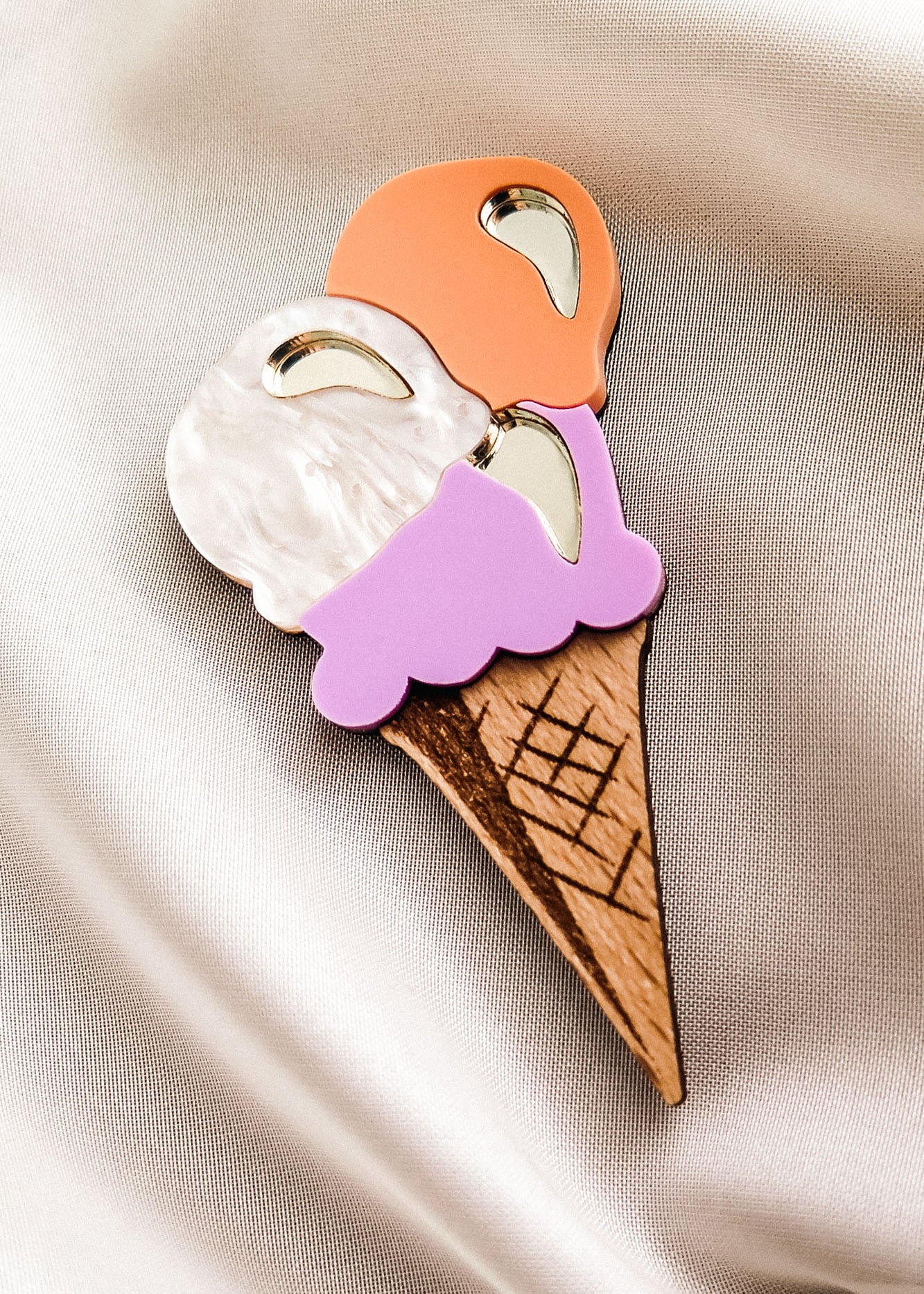 A handcrafted Ice Cream Brooch featuring three scoops—white, orange, and purple—on a wooden cone, displayed on smooth, light-colored satin fabric. Perfect for adding a playful touch to your summer look.
