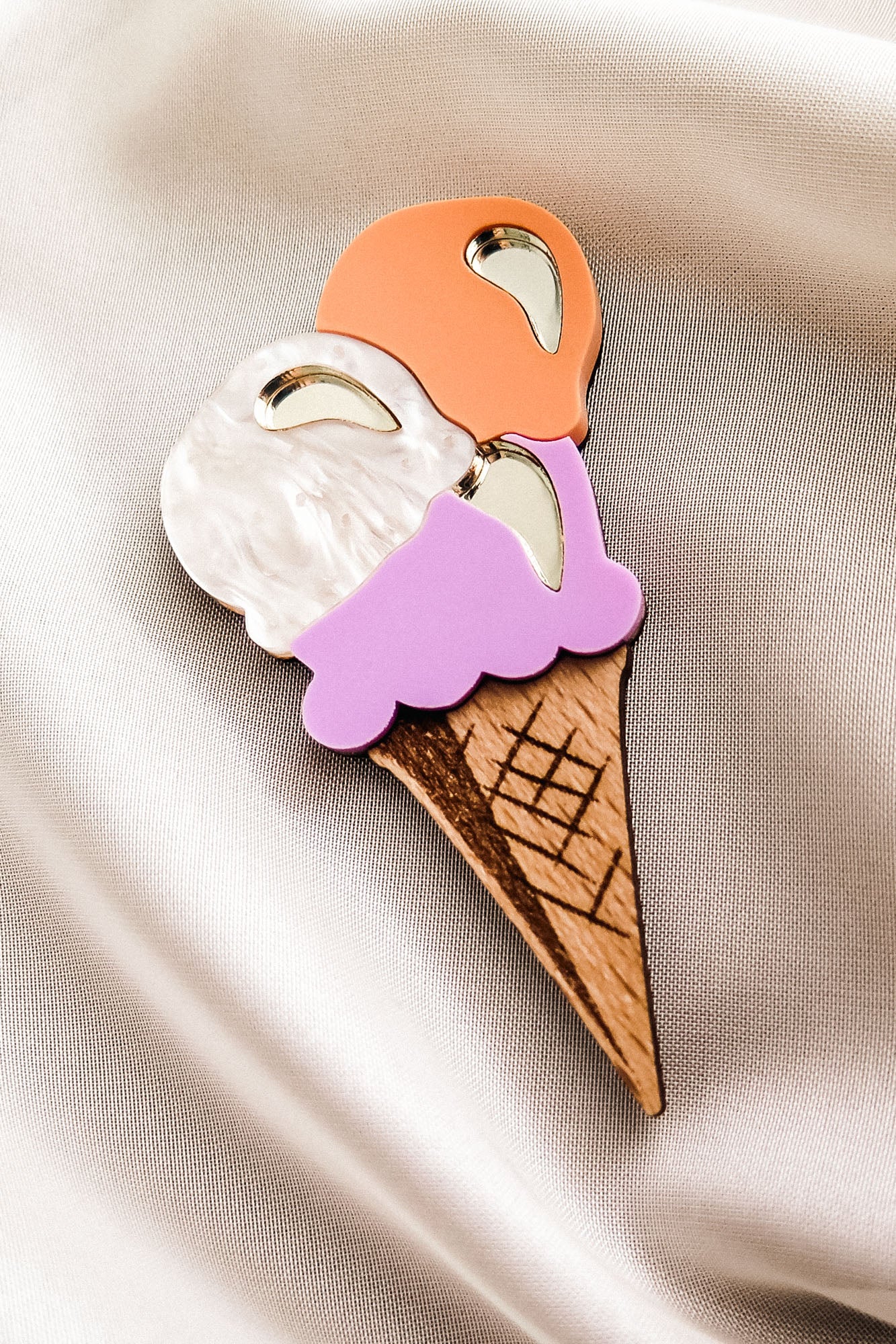 A handcrafted Ice Cream Brooch featuring three scoops—white, orange, and purple—on a wooden cone, displayed on smooth, light-colored satin fabric. Perfect for adding a playful touch to your summer look.