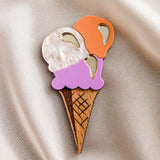 A handcrafted wooden ice cream brooch with white, orange, and purple scoops and shiny metallic accents is placed on light beige satin fabric—perfect for elevating your summer look.