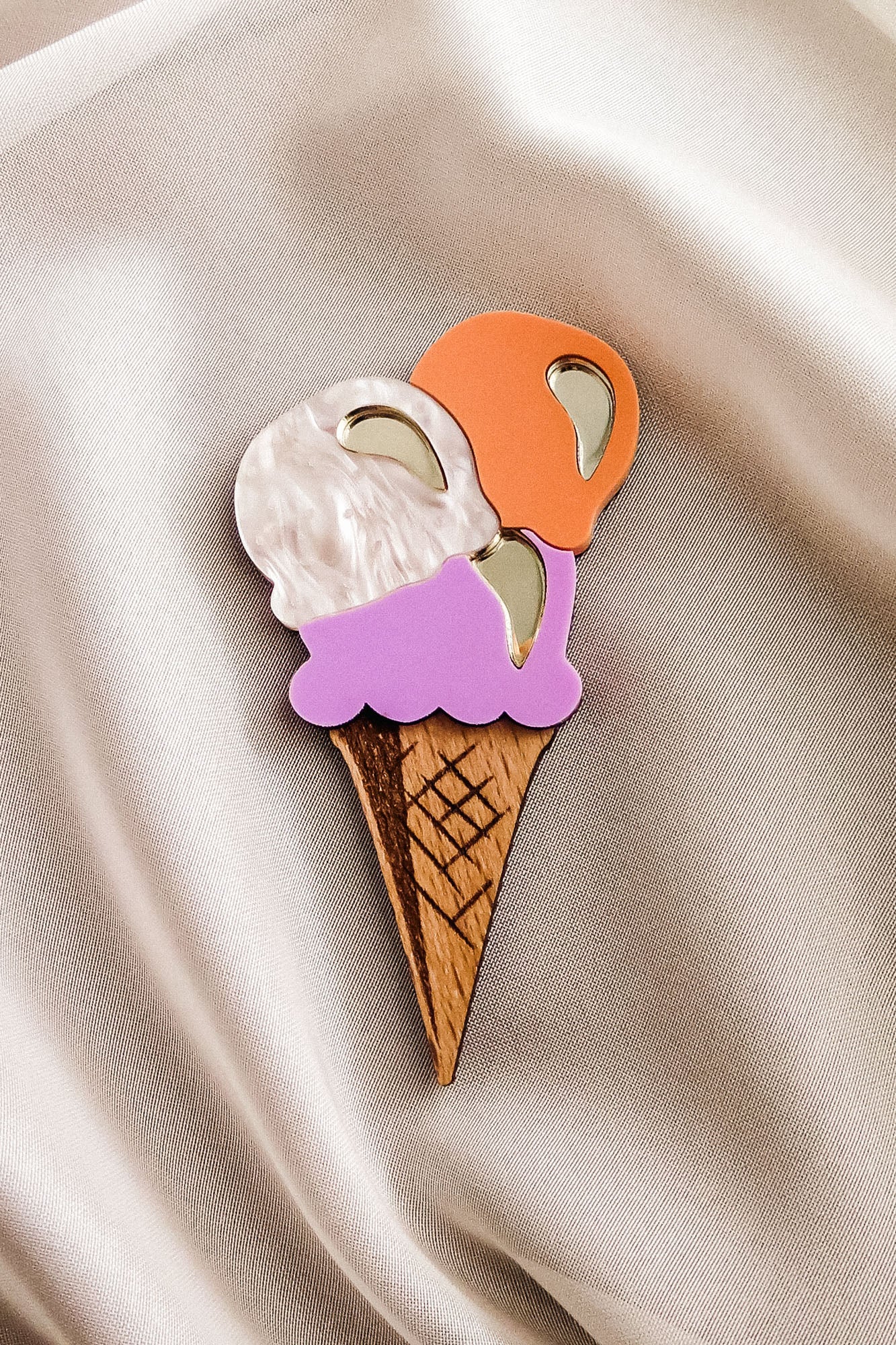 A handcrafted wooden ice cream brooch with white, orange, and purple scoops and shiny metallic accents is placed on light beige satin fabric—perfect for elevating your summer look.