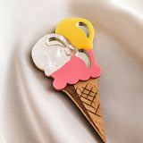 A handcrafted accessory, this decorative wooden Ice Cream Brooch with three scoops in yellow, white, and pink rests on smooth, cream-colored fabric—perfect for brightening up your summer look.