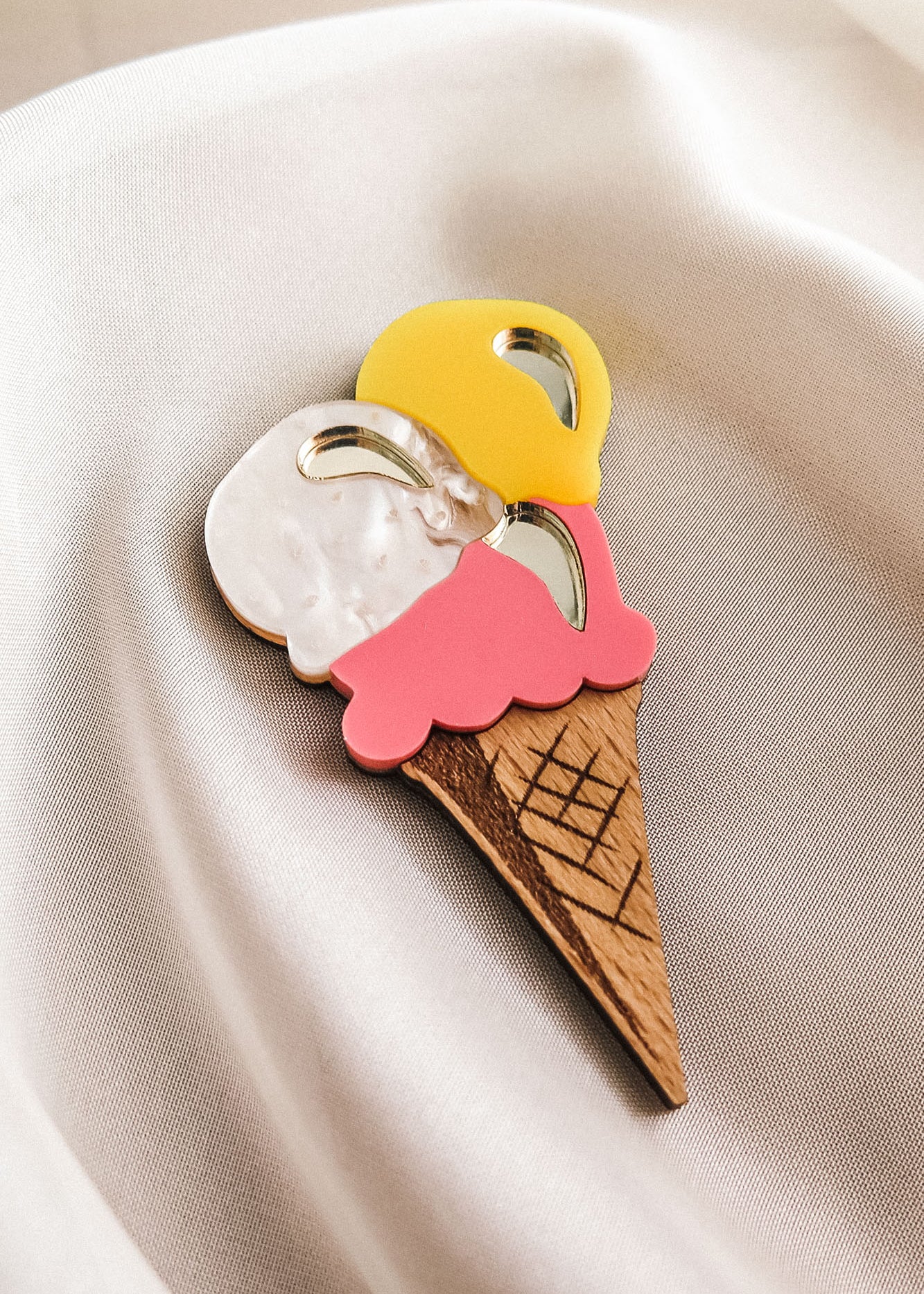 A handcrafted accessory, this decorative wooden Ice Cream Brooch with three scoops in yellow, white, and pink rests on smooth, cream-colored fabric—perfect for brightening up your summer look.