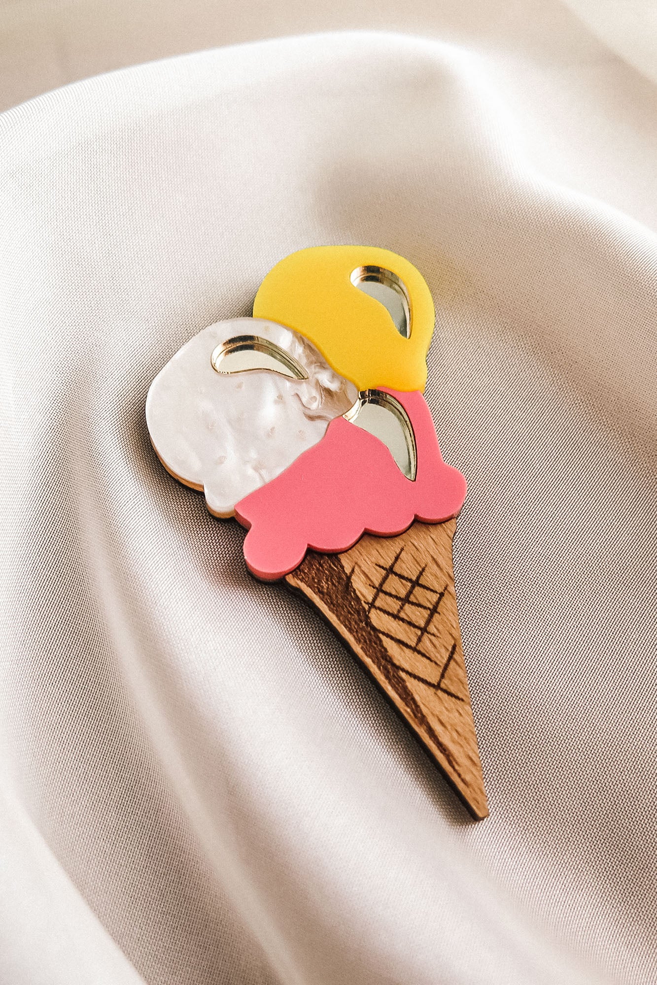 A handcrafted accessory, this decorative wooden Ice Cream Brooch with three scoops in yellow, white, and pink rests on smooth, cream-colored fabric—perfect for brightening up your summer look.