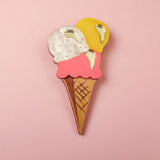 A handcrafted accessory, this wooden Ice Cream Brooch features three scoops—white, pink, and yellow—with shiny gold accents, perfect for adding a playful touch to your summer look. Displayed on a pink background.