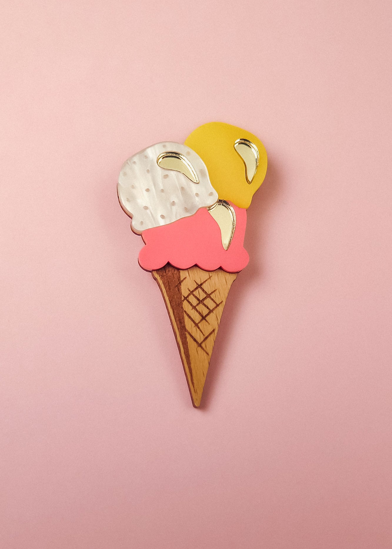 A handcrafted accessory, this wooden Ice Cream Brooch features three scoops—white, pink, and yellow—with shiny gold accents, perfect for adding a playful touch to your summer look. Displayed on a pink background.