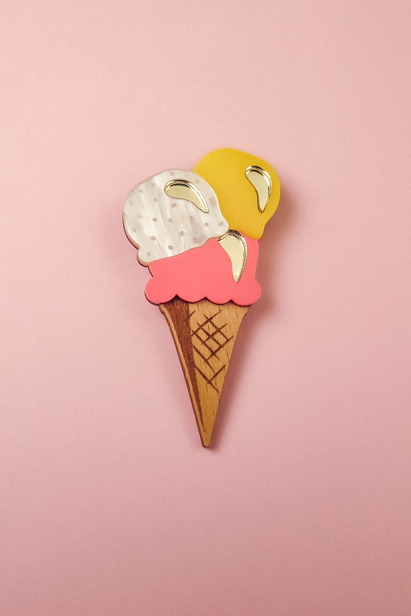 A handcrafted accessory, this wooden Ice Cream Brooch features three scoops—white, pink, and yellow—with shiny gold accents, perfect for adding a playful touch to your summer look. Displayed on a pink background.
