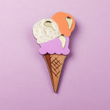 Handcrafted for a fun summer look, this Ice Cream Brooch features a wooden cone with three gold-accented scoops—white, purple, and orange—set against a pastel purple background.