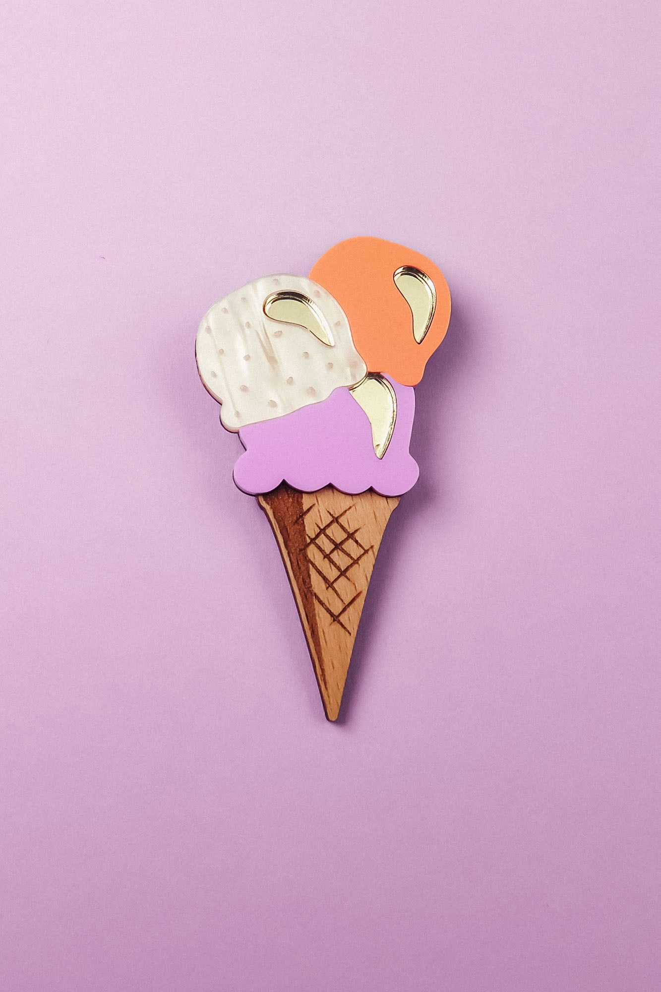 Handcrafted for a fun summer look, this Ice Cream Brooch features a wooden cone with three gold-accented scoops—white, purple, and orange—set against a pastel purple background.