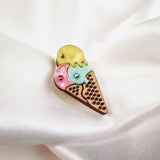 A whimsical handcrafted Ice Cream Pin features yellow, blue, and pink scoops topped with rhinestones on a textured cone, displayed on soft white fabric. This playful design adds charm to any outfit.