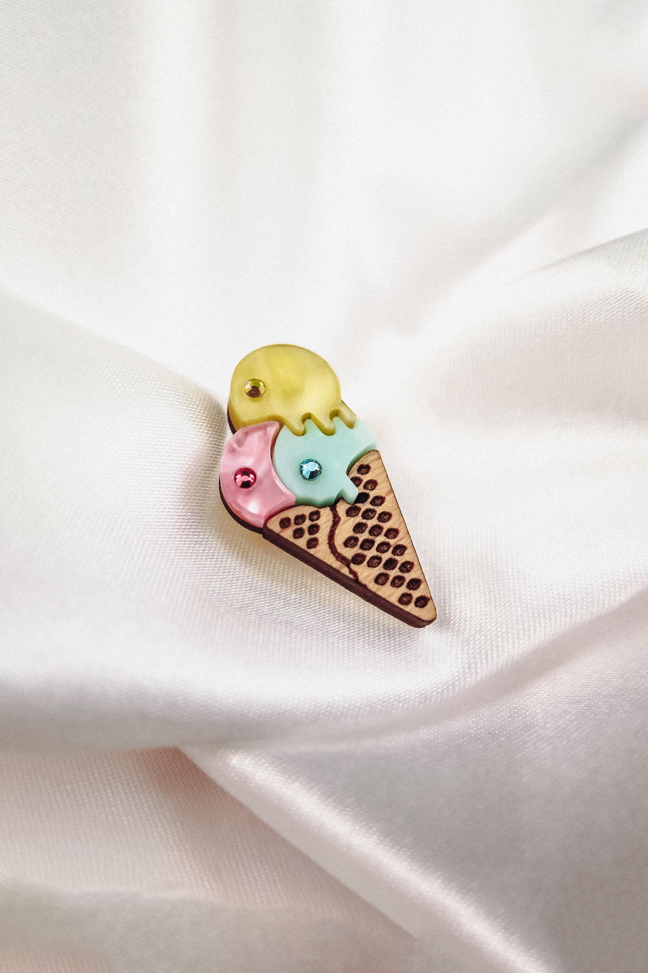 A whimsical handcrafted Ice Cream Pin features yellow, blue, and pink scoops topped with rhinestones on a textured cone, displayed on soft white fabric. This playful design adds charm to any outfit.