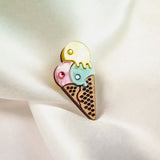 A handcrafted Ice Cream Pin featuring a colorful brooch with three scoops—yellow, pink, and blue—on a waffle cone, resting on soft white satin fabric. Its playful design adds charm to any outfit.