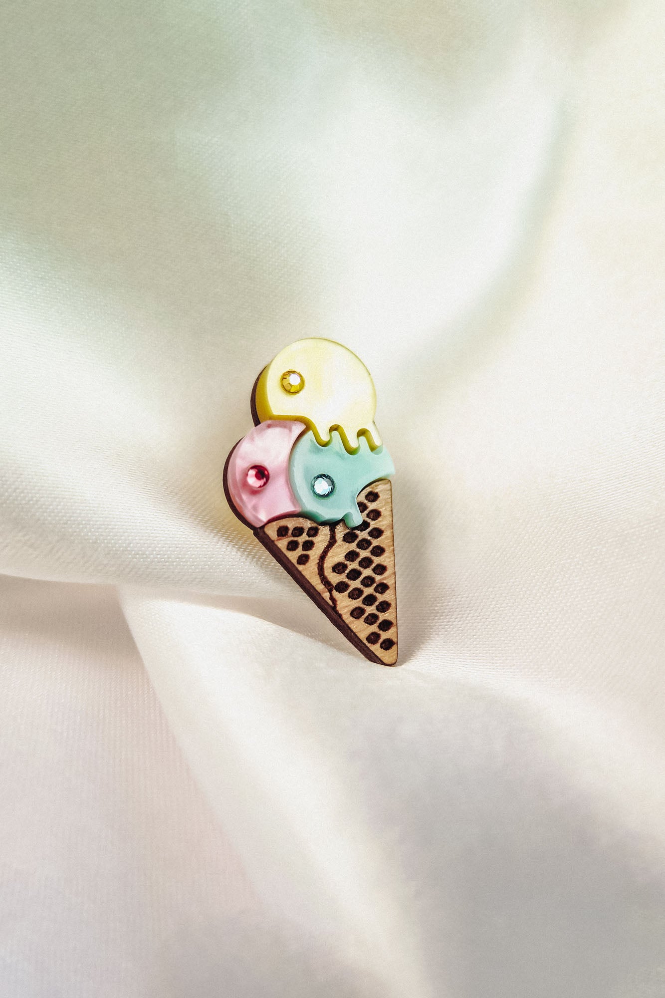 A handcrafted Ice Cream Pin featuring a colorful brooch with three scoops—yellow, pink, and blue—on a waffle cone, resting on soft white satin fabric. Its playful design adds charm to any outfit.