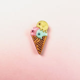 A playful design, this handcrafted Ice Cream Pin features a colorful enamel cone with three scoops—pink, blue, and yellow—set against a soft pink and white gradient background.