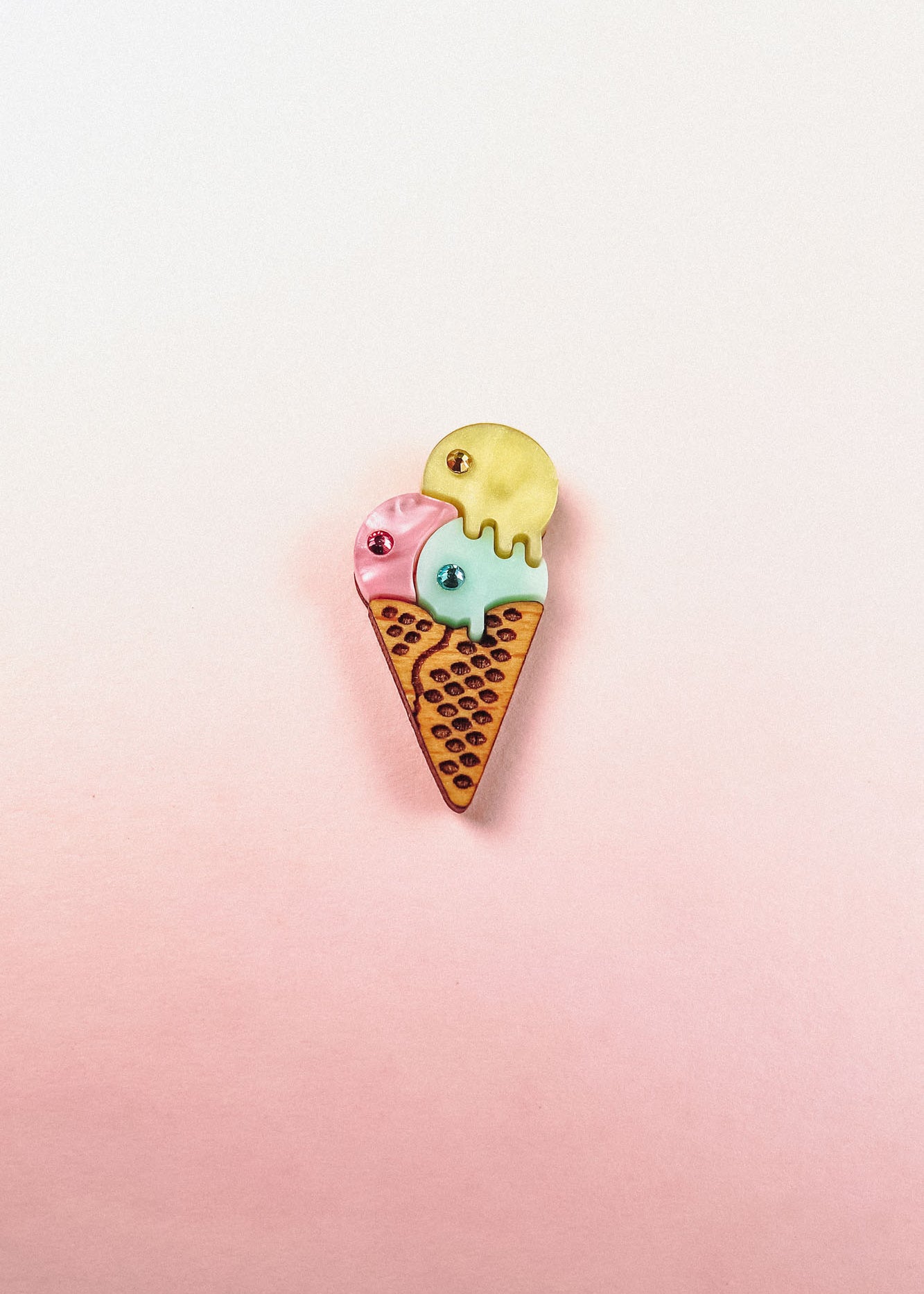 A playful design, this handcrafted Ice Cream Pin features a colorful enamel cone with three scoops—pink, blue, and yellow—set against a soft pink and white gradient background.