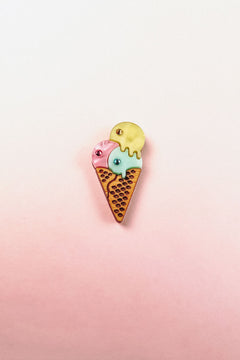 A playful design, this handcrafted Ice Cream Pin features a colorful enamel cone with three scoops—pink, blue, and yellow—set against a soft pink and white gradient background.