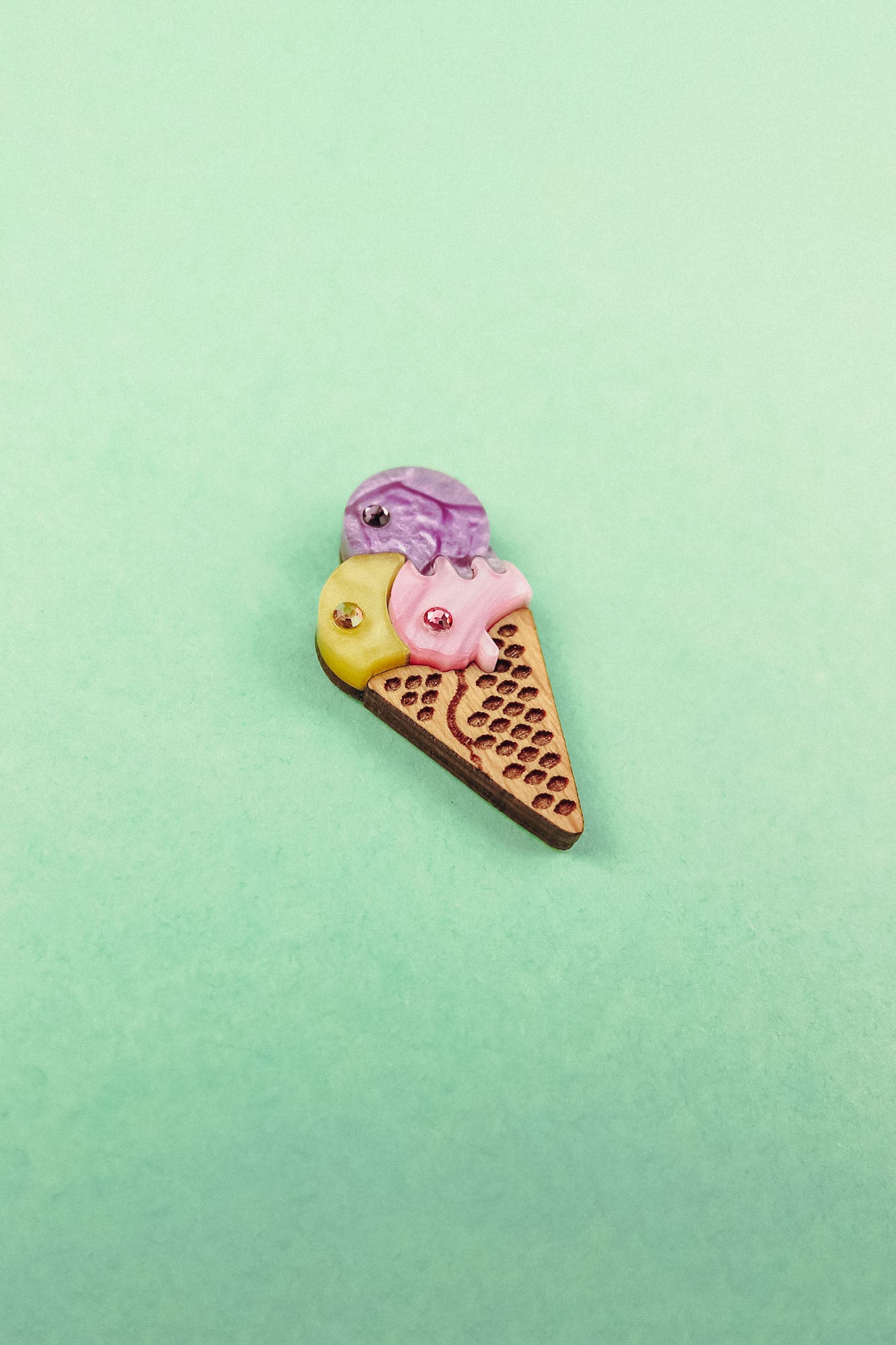 A small, handcrafted Ice Cream Pin with a playful design featuring yellow, purple, and pink scoops topped with gemstone accents, set against a pastel green background.