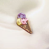 A handcrafted Ice Cream Pin shaped like a cone with three scoops—yellow, pink, and purple—features a playful design with small embedded rhinestones, resting on a soft, light-colored fabric background.