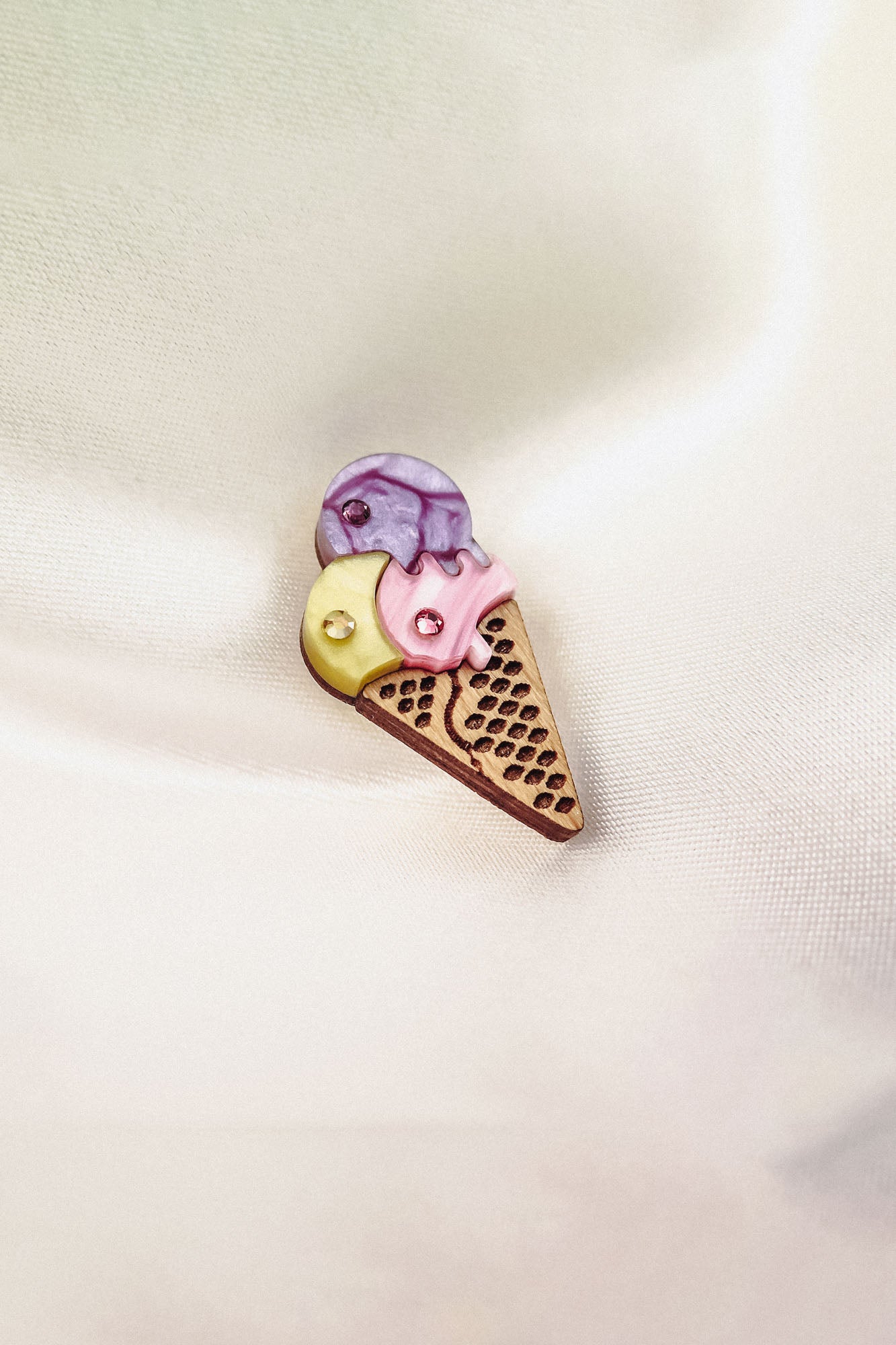 A handcrafted Ice Cream Pin shaped like a cone with three scoops—yellow, pink, and purple—features a playful design with small embedded rhinestones, resting on a soft, light-colored fabric background.