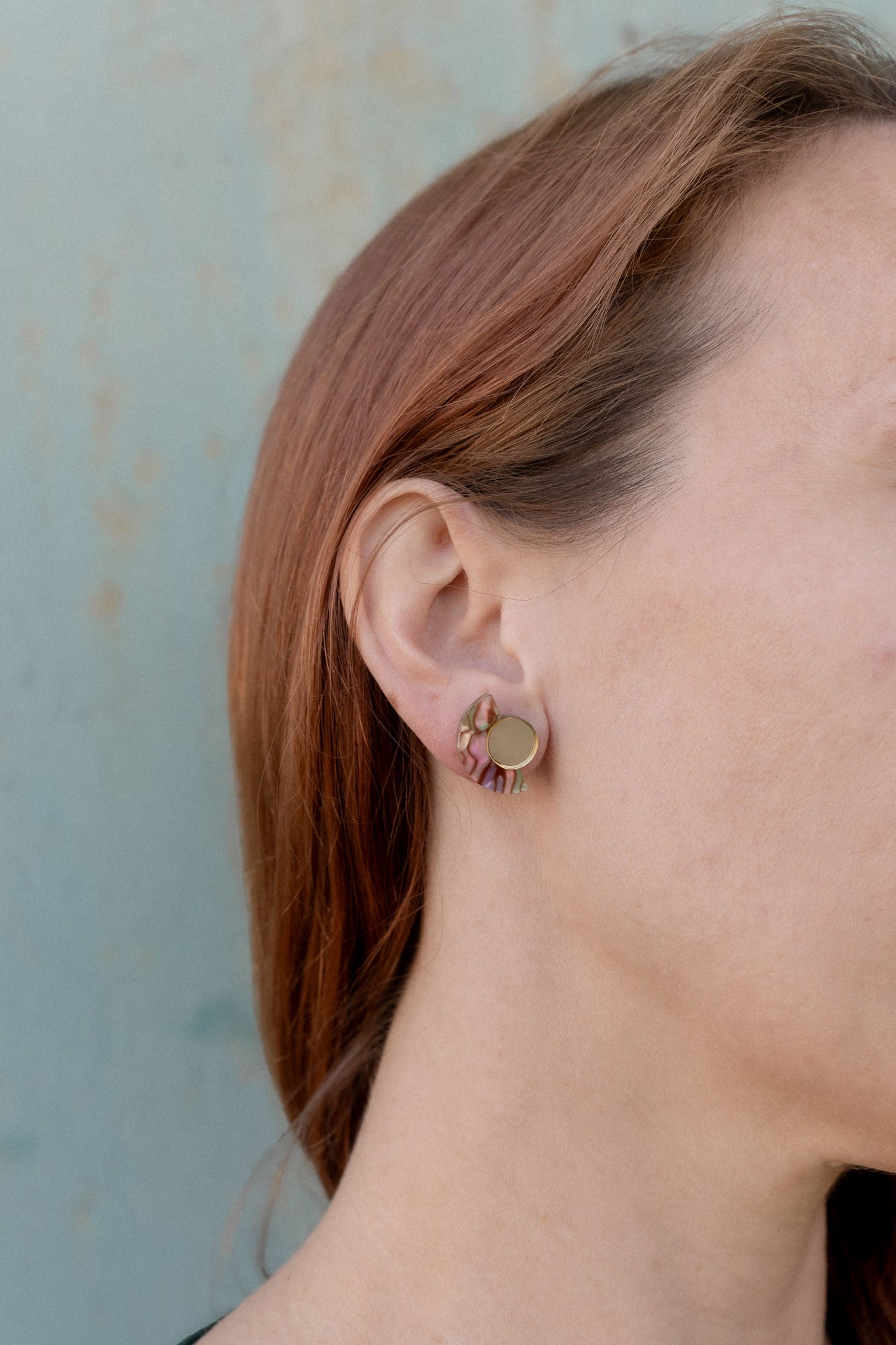 Close-up of a person with light skin and reddish-brown hair wearing a small, round, gold Minimalist Geometric Stud Earring featuring a wavy, pink and green patterned design on their left ear against a light blue background.