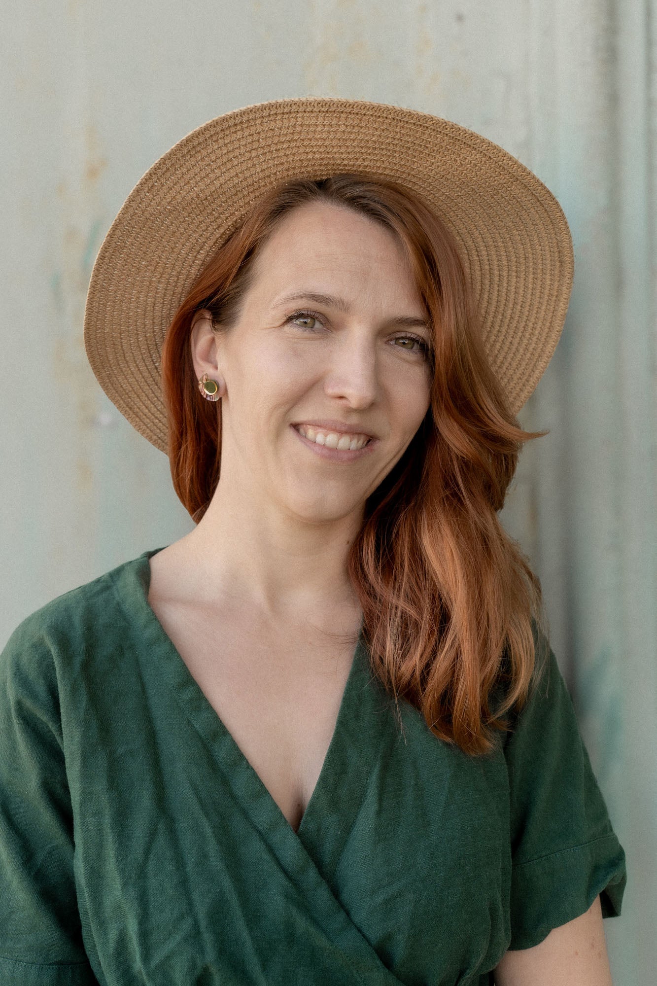 A woman with long auburn hair wears minimalist geometric stud earrings, a wide-brimmed straw hat, and a green dress, smiling softly while standing in front of a neutral-colored wall.