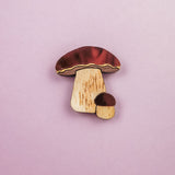 A wooden King Bolete Mushroom Pin, featuring brown caps and light stems, showcases handcrafted quality and a nature-inspired design against a pastel pink background.