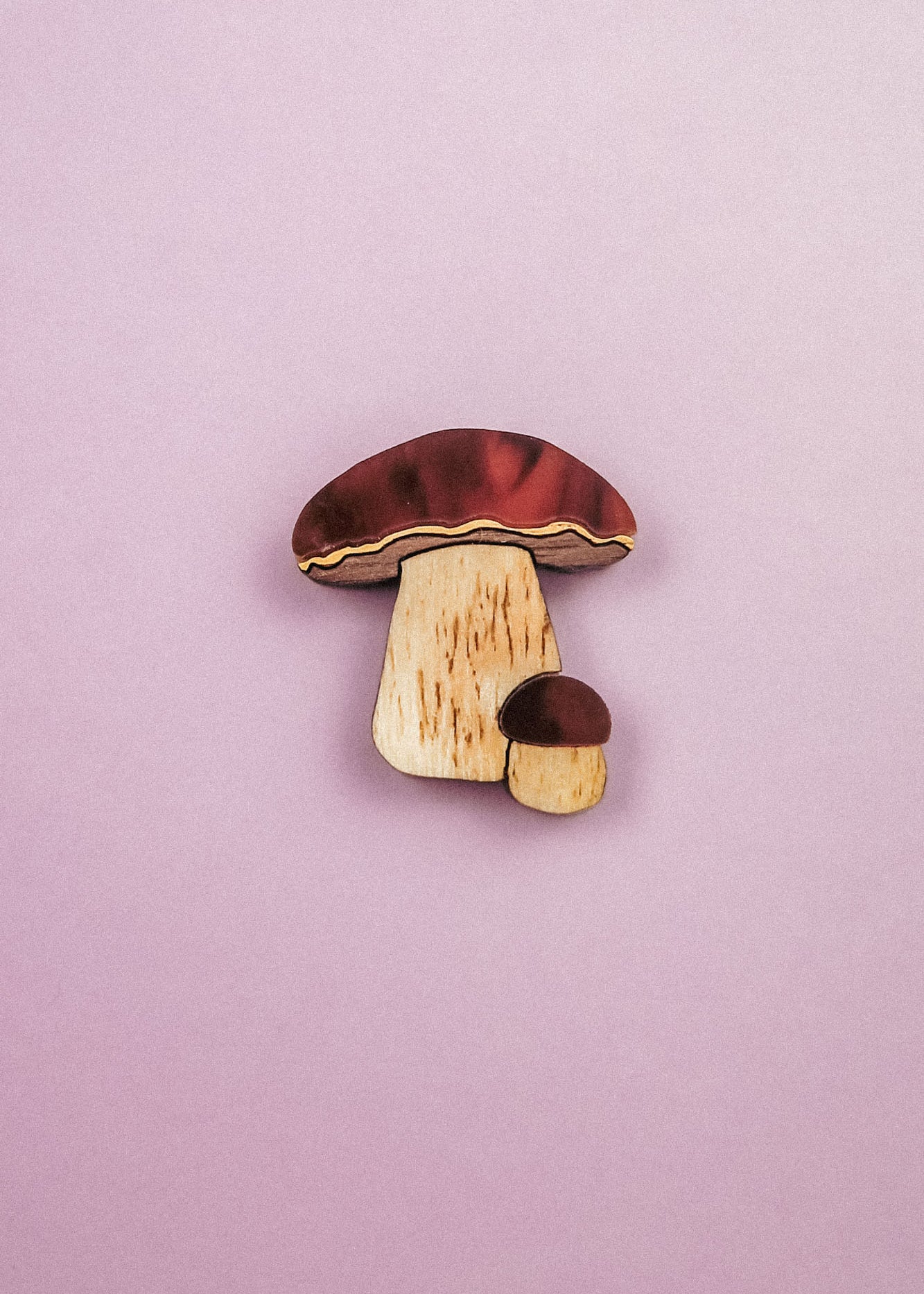A wooden King Bolete Mushroom Pin, featuring brown caps and light stems, showcases handcrafted quality and a nature-inspired design against a pastel pink background.