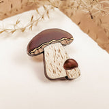 A wooden brooch featuring a nature-inspired design of two brown-capped mushrooms, one large and one small, placed on white paper with dried beige plants in the background. Showcases handcrafted quality and resembles a King Bolete Mushroom Pin.