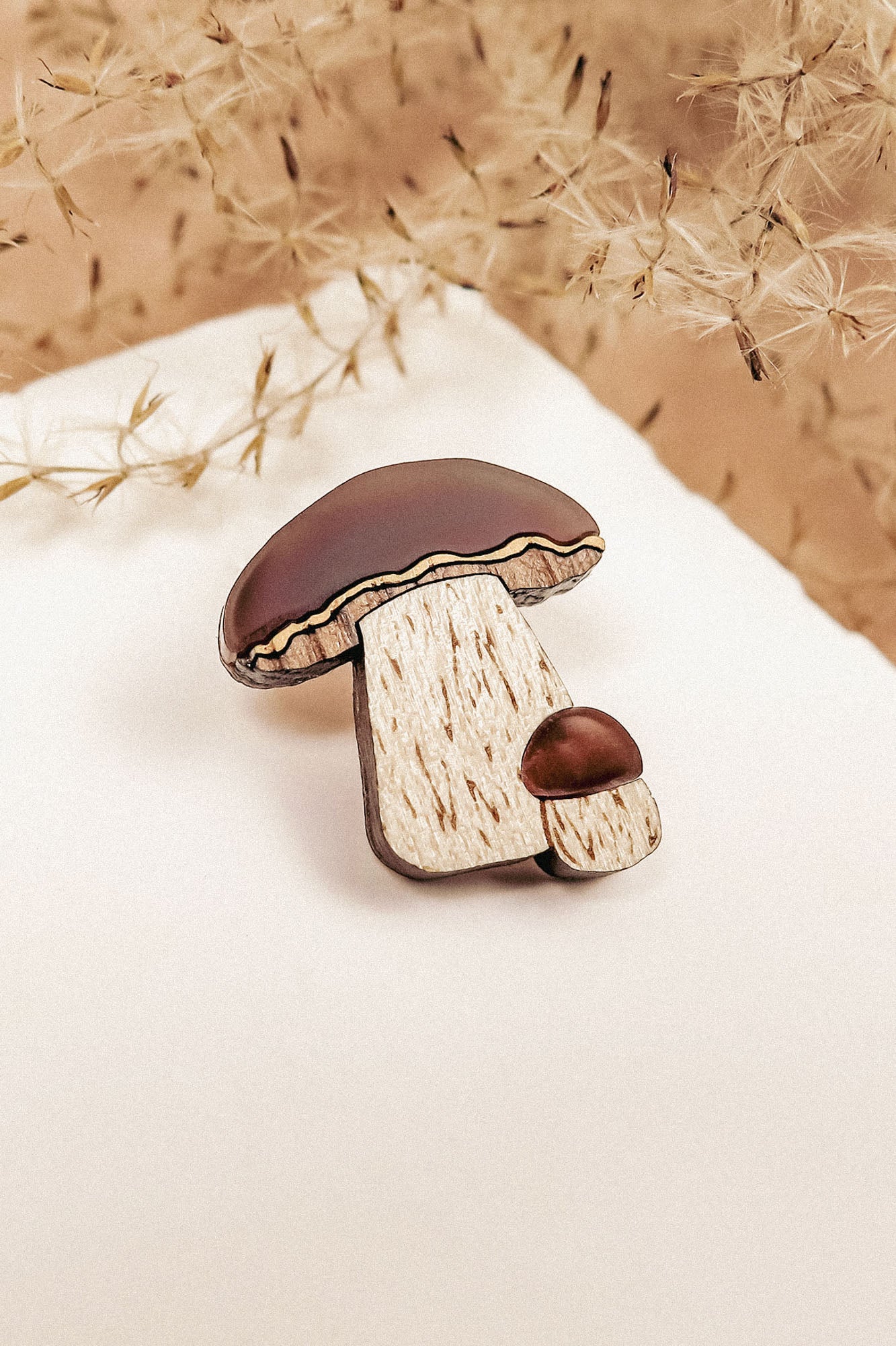 A wooden brooch featuring a nature-inspired design of two brown-capped mushrooms, one large and one small, placed on white paper with dried beige plants in the background. Showcases handcrafted quality and resembles a King Bolete Mushroom Pin.