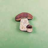 A nature-inspired design, this King Bolete Mushroom Pin features handcrafted quality with two mushrooms—one large and one small—sporting brown caps and beige stems, set against a textured green background.