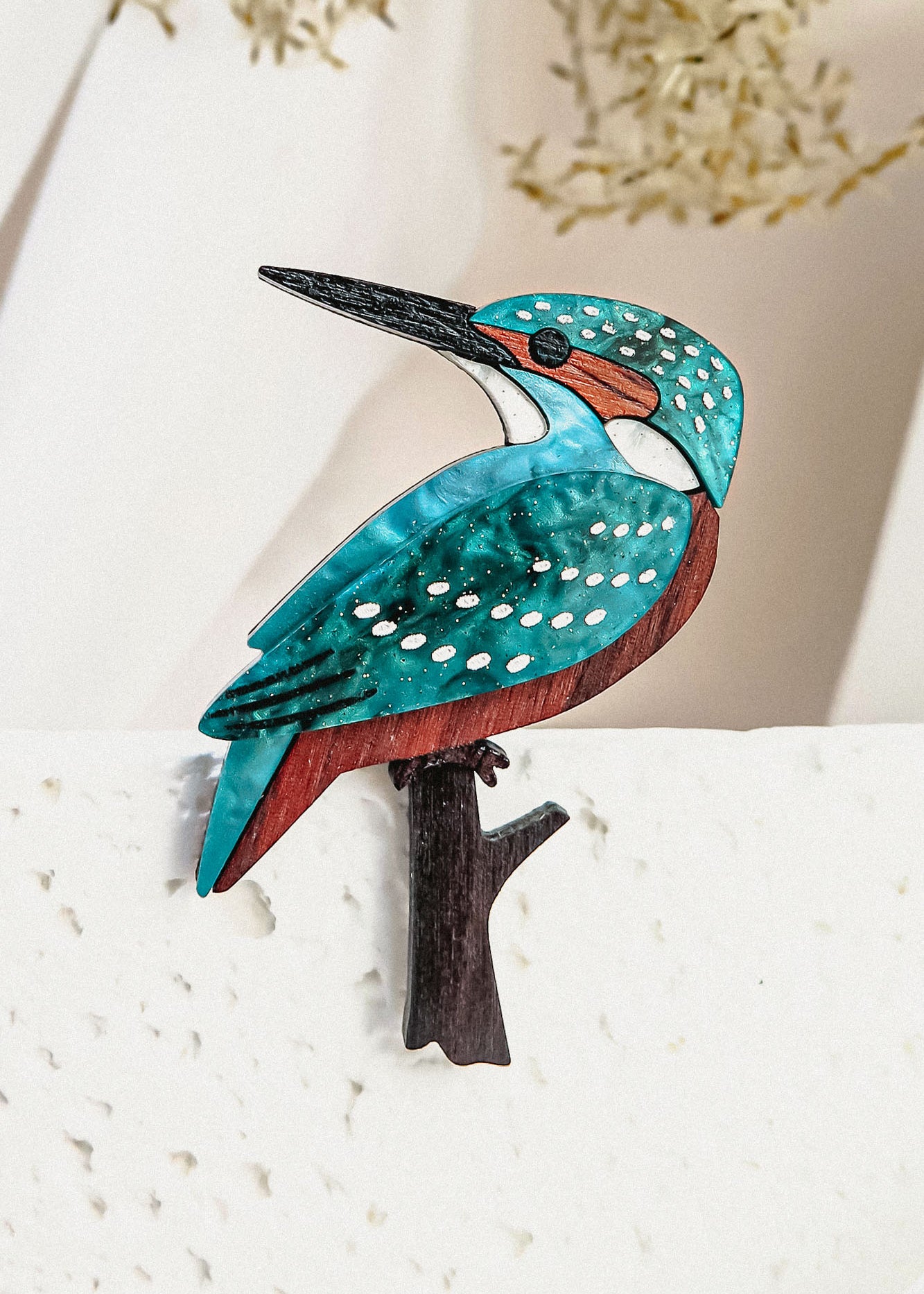 A colorful handcrafted Kingfisher Brooch featuring turquoise, brown, and white details perches on a textured white surface, with blurred beige foliage in the background.