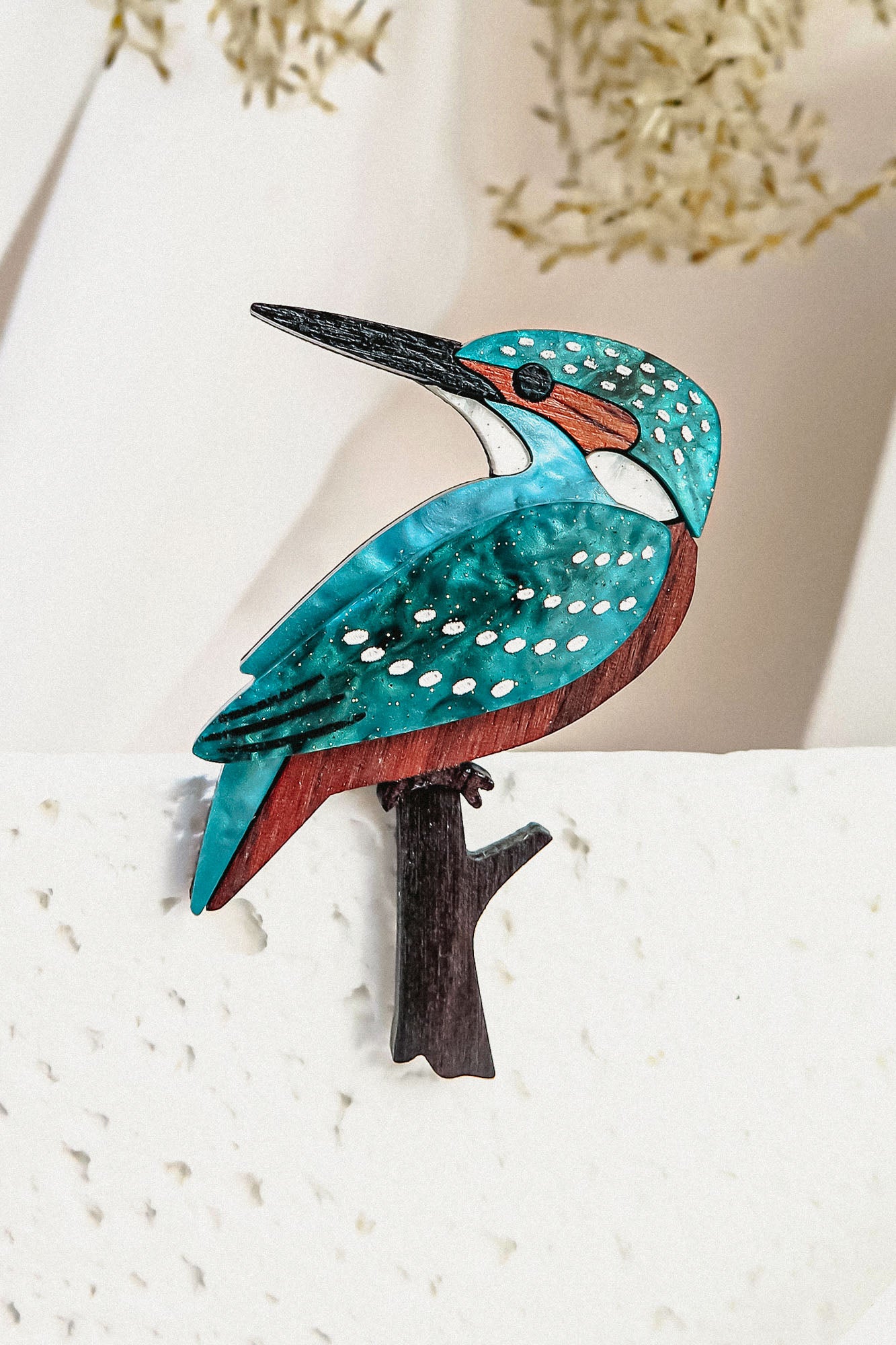 A colorful handcrafted Kingfisher Brooch featuring turquoise, brown, and white details perches on a textured white surface, with blurred beige foliage in the background.