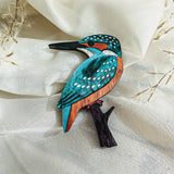 A handcrafted Kingfisher Brooch made from sustainable materials, painted in teal, orange, and white, rests on cream-colored fabric with delicate dried grass in the background.