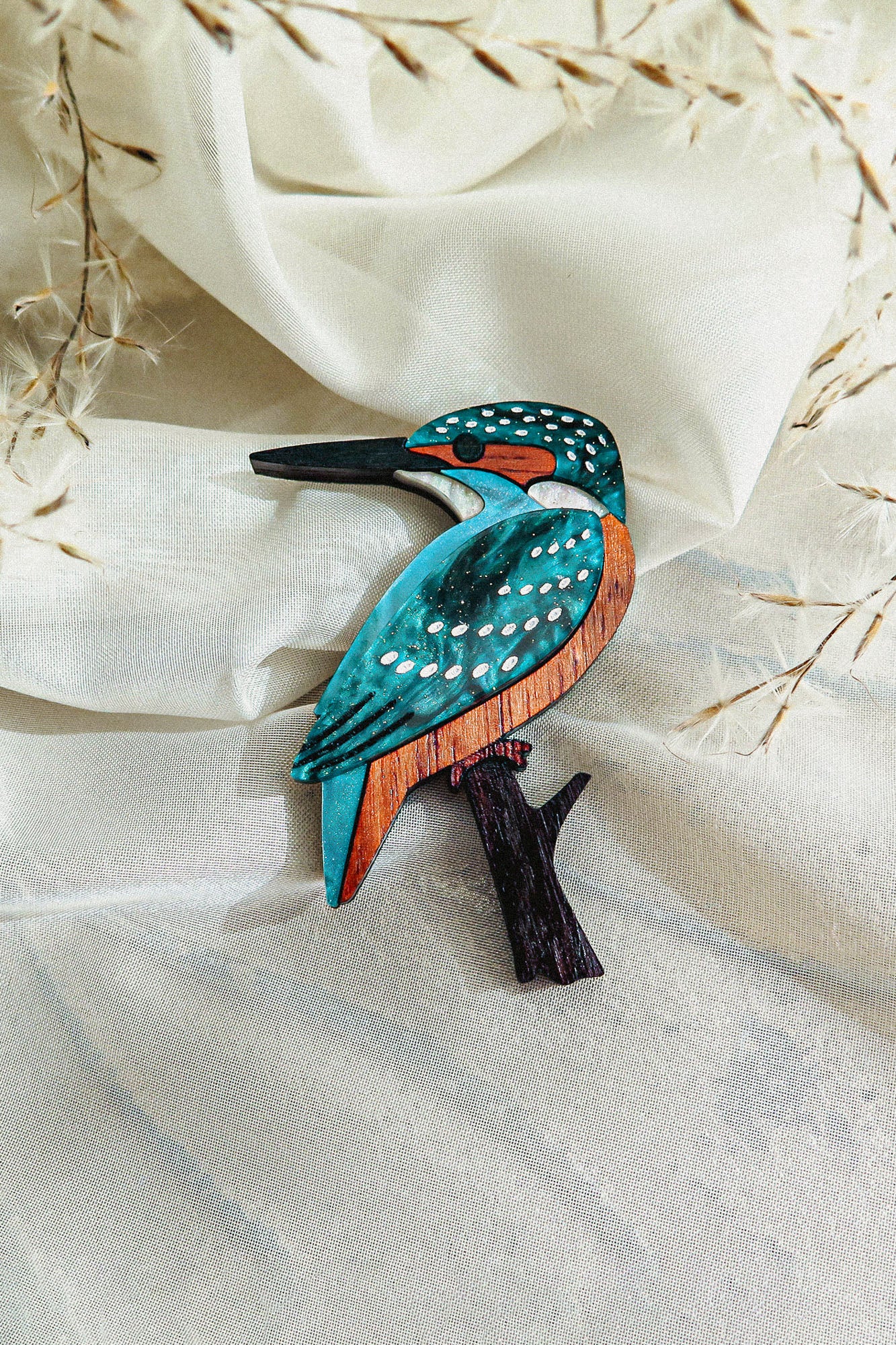 A handcrafted Kingfisher Brooch made from sustainable materials, painted in teal, orange, and white, rests on cream-colored fabric with delicate dried grass in the background.