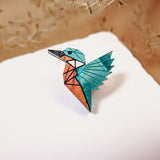A colorful origami bird crafted from eco-friendly materials with blue, teal, and orange sections sits on a white surface, delicate dried grass or dandelion seeds softly blurred in the background.