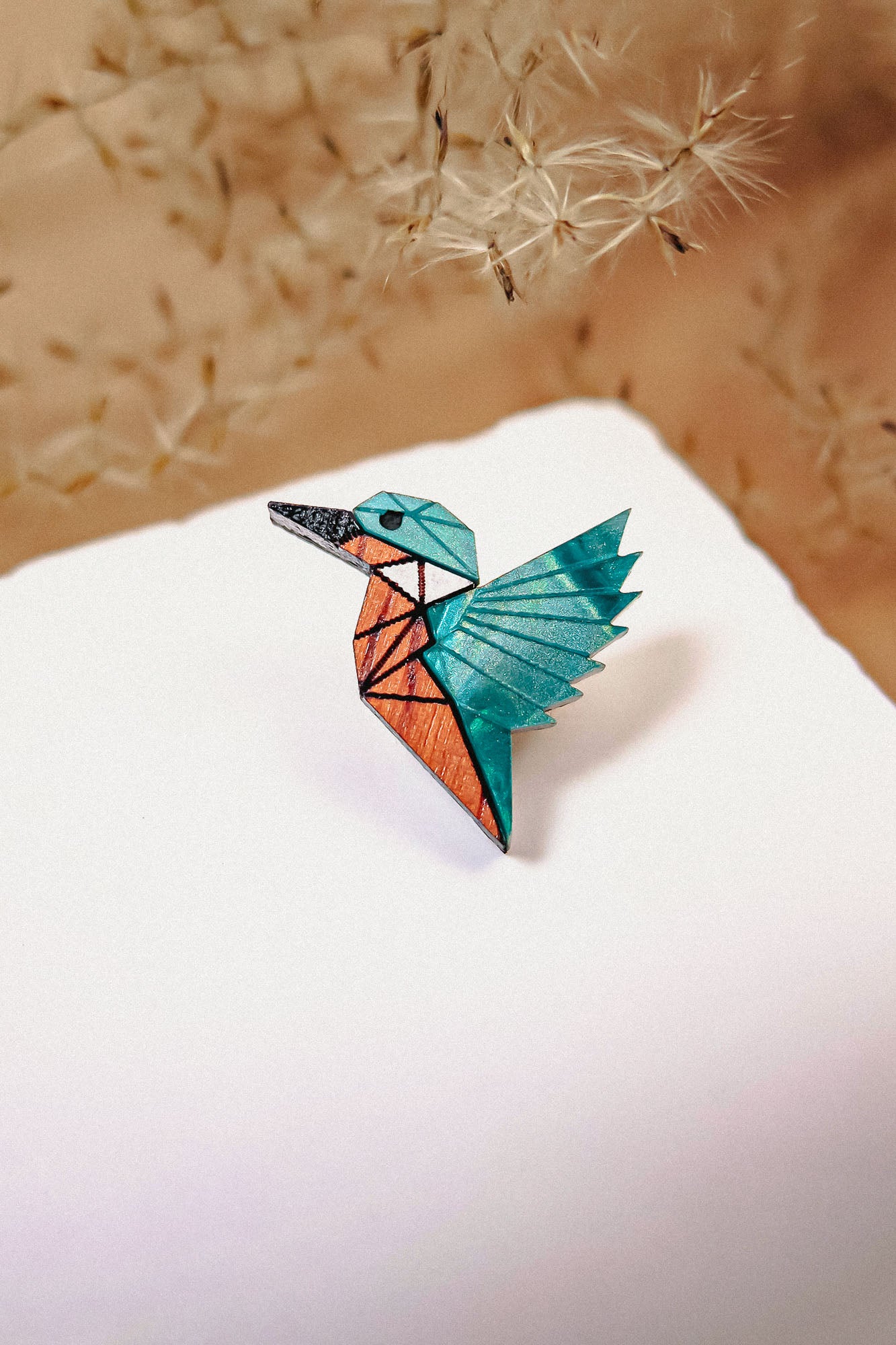 A colorful origami bird crafted from eco-friendly materials with blue, teal, and orange sections sits on a white surface, delicate dried grass or dandelion seeds softly blurred in the background.