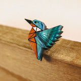 A colorful origami-style kingfisher pin brooch with blue, teal, and orange geometric sections, perched on the edge of recycled wood against a soft beige background—crafted from eco-friendly materials.