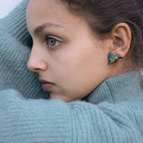 A young person wearing a teal knit sweater looks to the side, showing a Kingfisher Stud Earring. Their hair is pulled back, and their expression appears calm and thoughtful, embracing an eco-friendly, sustainably made accessory.