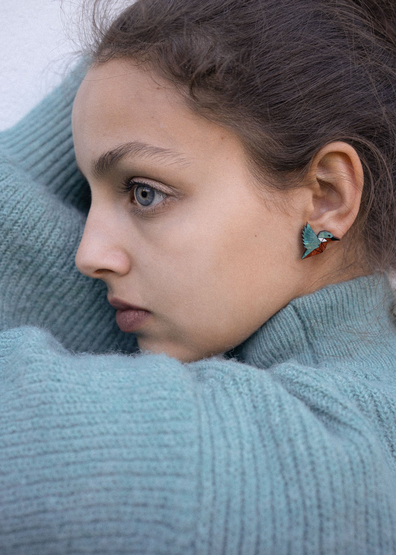 A young person wearing a teal knit sweater looks to the side, showing a Kingfisher Stud Earring. Their hair is pulled back, and their expression appears calm and thoughtful, embracing an eco-friendly, sustainably made accessory.