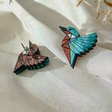 Sustainably made Kingfisher Stud Earrings, one painted in blue, white, and brown geometric patterns and the other left natural wood, rest on light sheer fabric with soft dried plant stems above them.