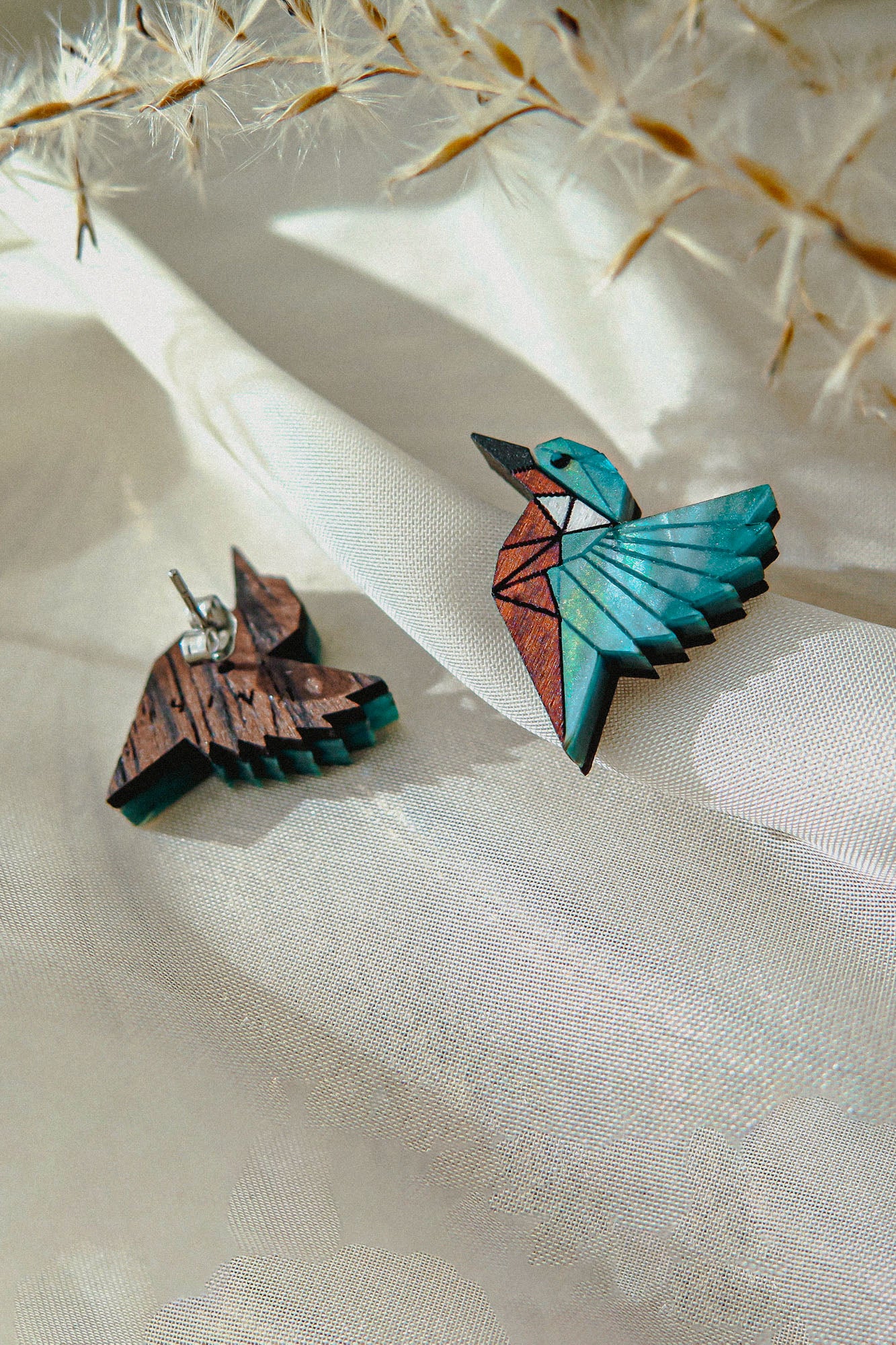 Sustainably made Kingfisher Stud Earrings, one painted in blue, white, and brown geometric patterns and the other left natural wood, rest on light sheer fabric with soft dried plant stems above them.