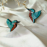 Two colorful, geometric bird brooches, sustainably made and resembling hummingbirds, rest on soft white fabric, with delicate dried plant stems in the background.