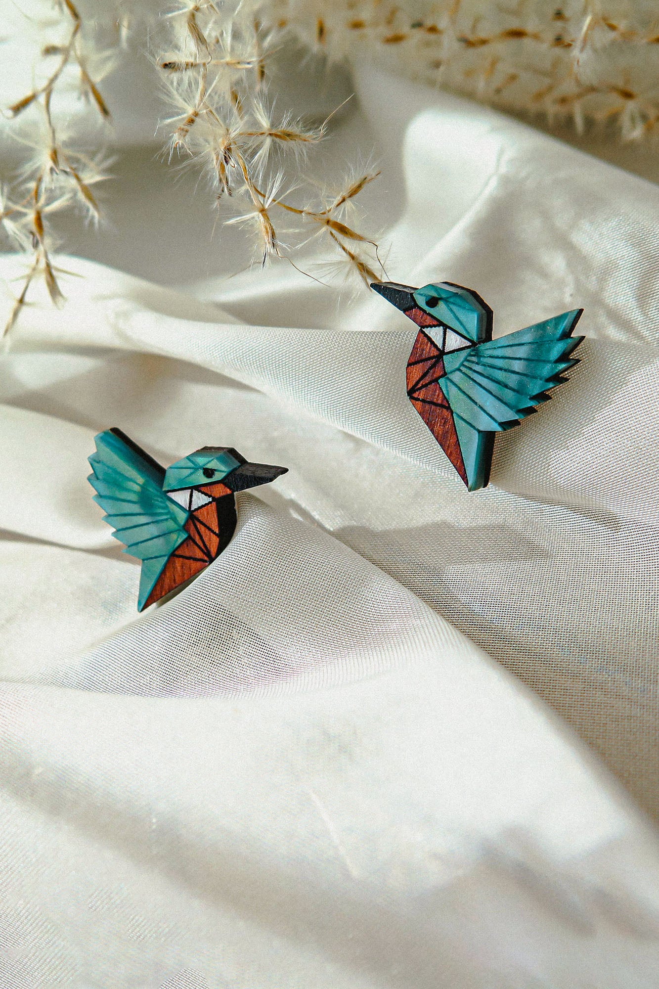Two colorful, geometric bird brooches, sustainably made and resembling hummingbirds, rest on soft white fabric, with delicate dried plant stems in the background.
