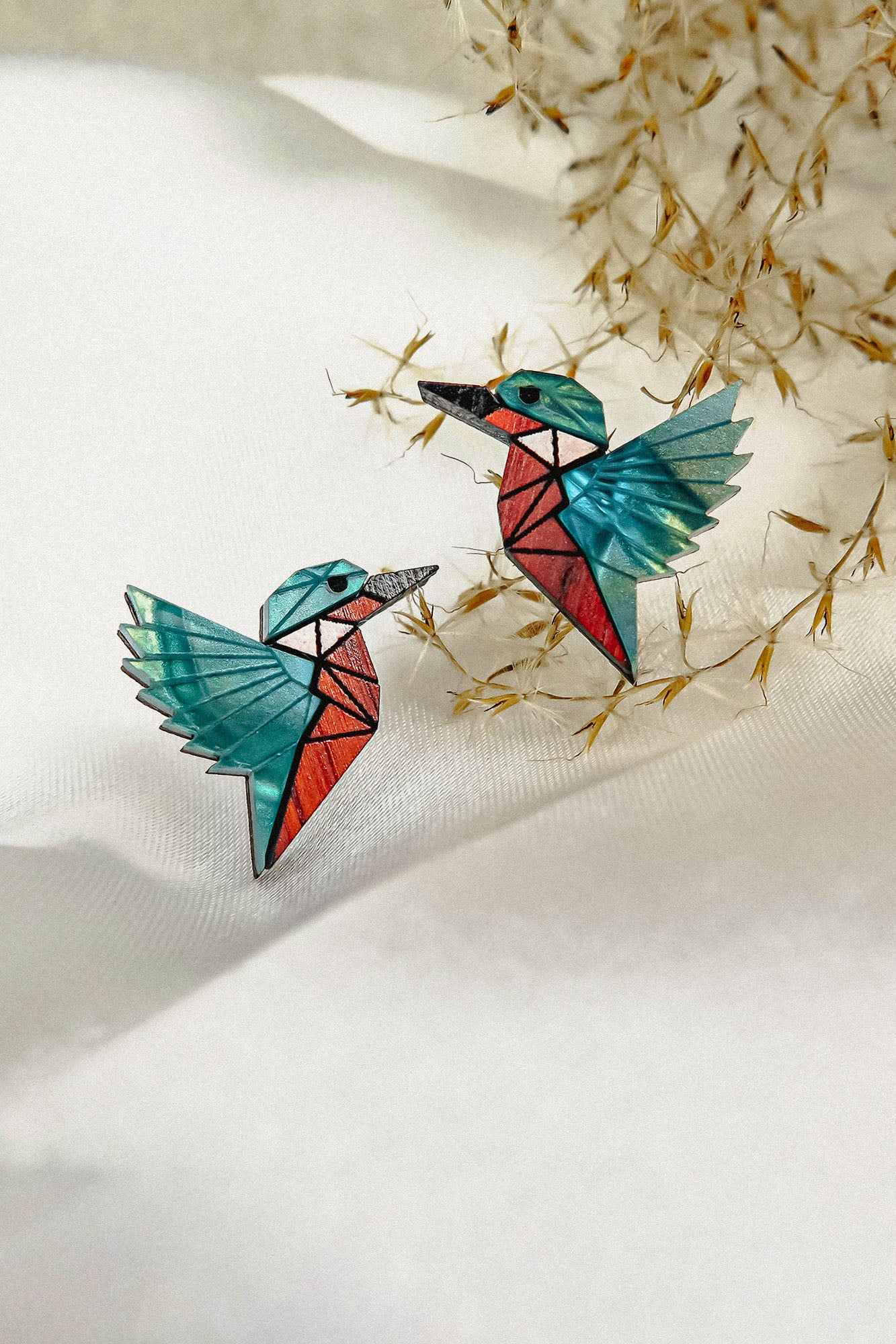 Eco-friendly Kingfisher Stud Earrings: Two colorful origami-style bird earrings with turquoise wings and red bodies, placed on a white textured surface with delicate dried grass in the background.