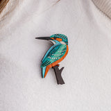 A handcrafted Kingfisher Brooch, featuring blue, orange, and white details, is pinned to a white textured sweater. Strands of brown hair are partially visible at the top of the image. Made using sustainable materials.