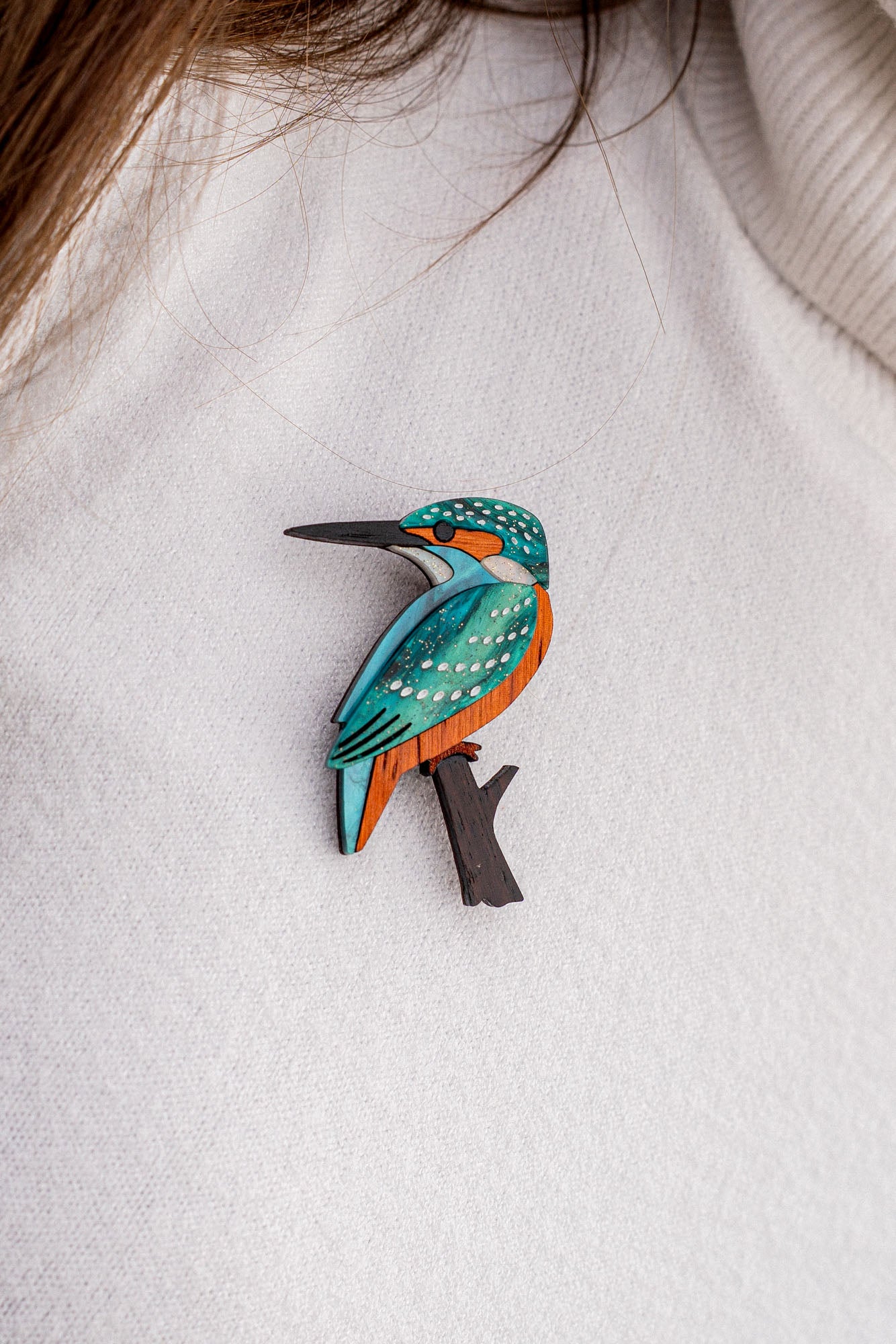 A handcrafted Kingfisher Brooch, featuring blue, orange, and white details, is pinned to a white textured sweater. Strands of brown hair are partially visible at the top of the image. Made using sustainable materials.