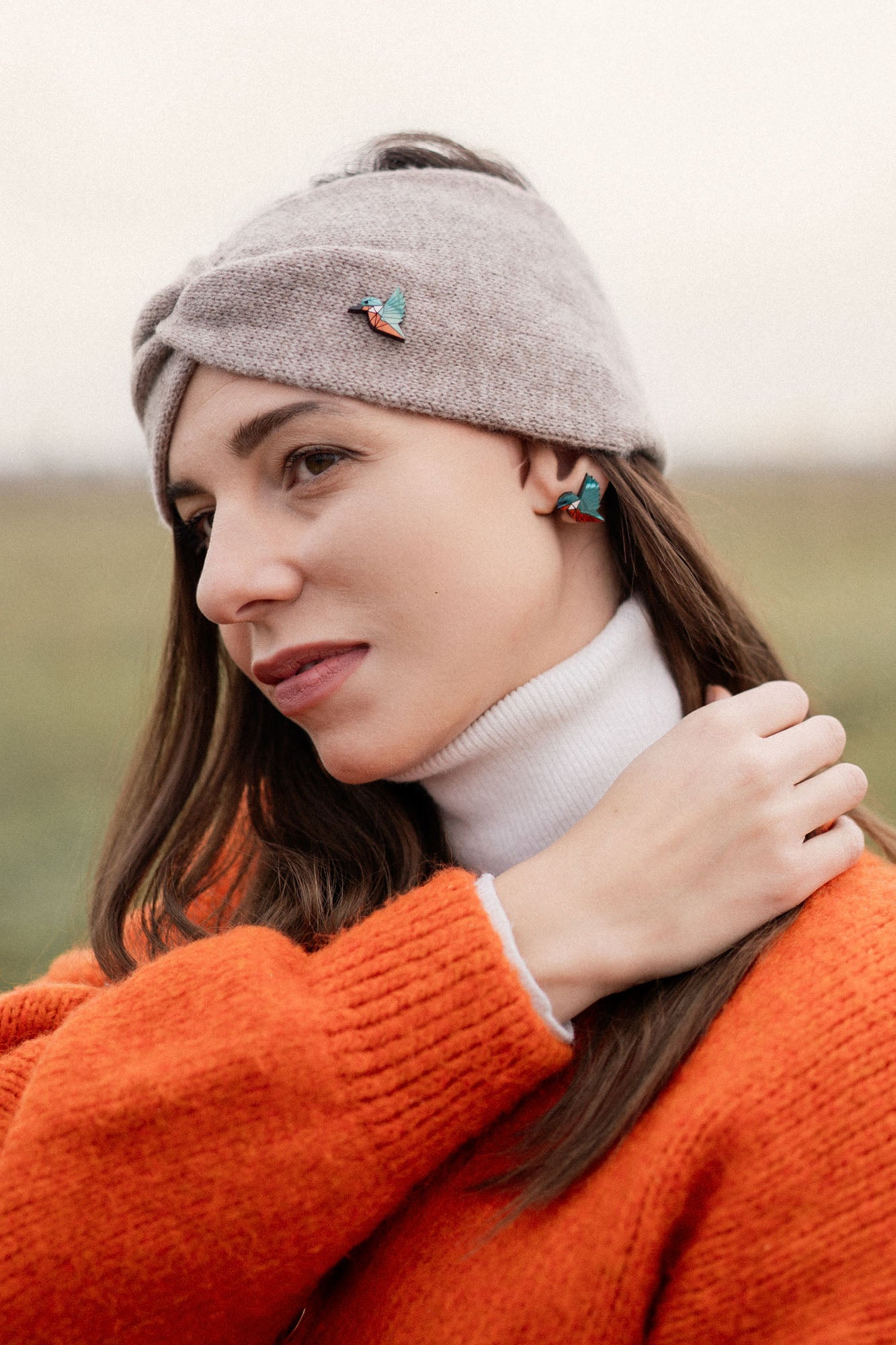 A woman wearing a grey headband with a kingfisher pin, a white turtleneck, and an orange sweater stands outdoors, looking to the side and touching her hair. The background is blurred and grassy—perfect for bird lovers seeking an eco-friendly accessory.
